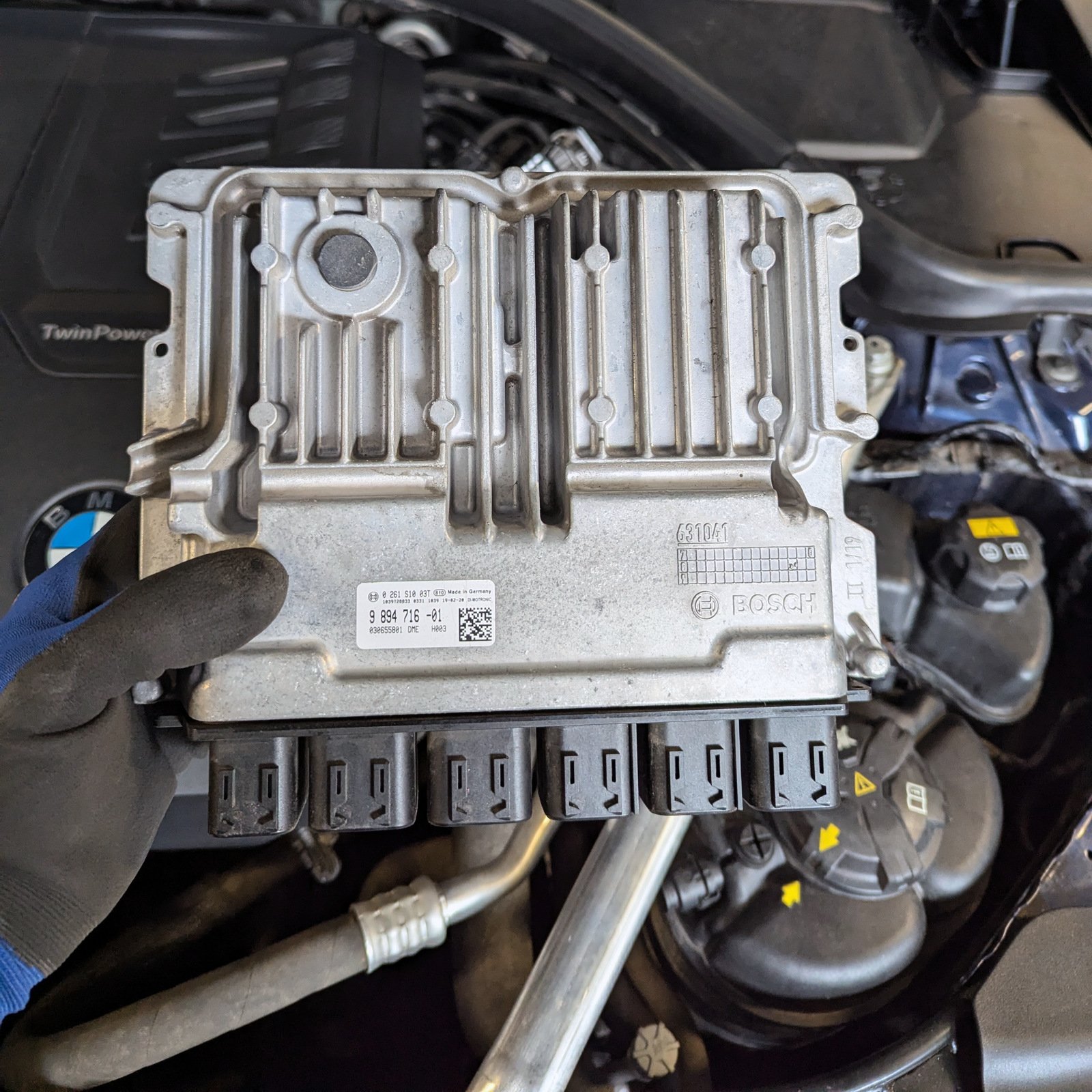 Bosch DME engine control unit held in a gloved hand above the BMW engine bay, showing the metal housing and six connectors.