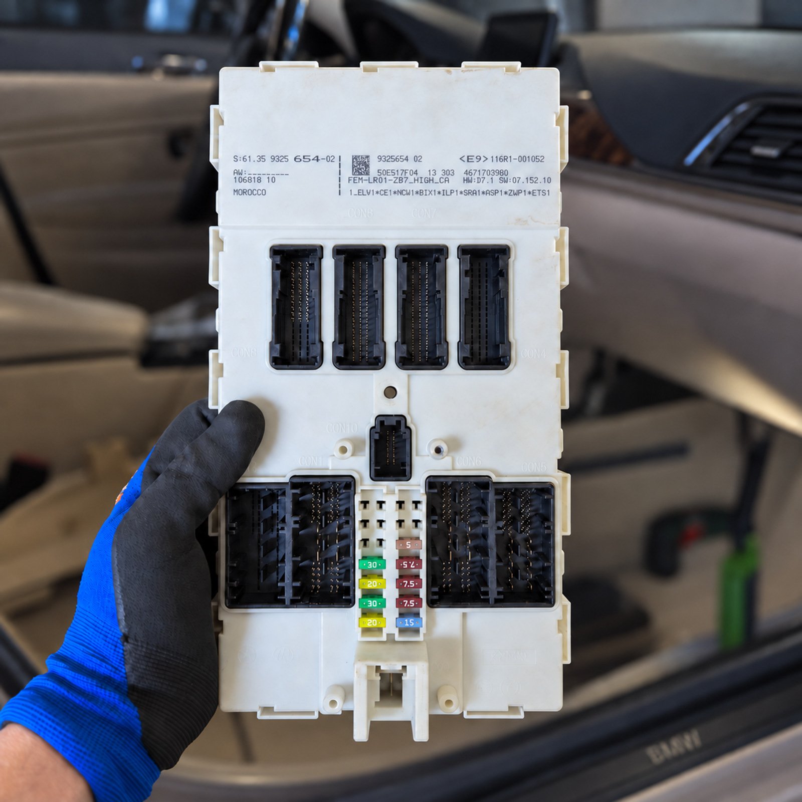 BMW FEM control module held in a gloved hand beside the vehicle interior
