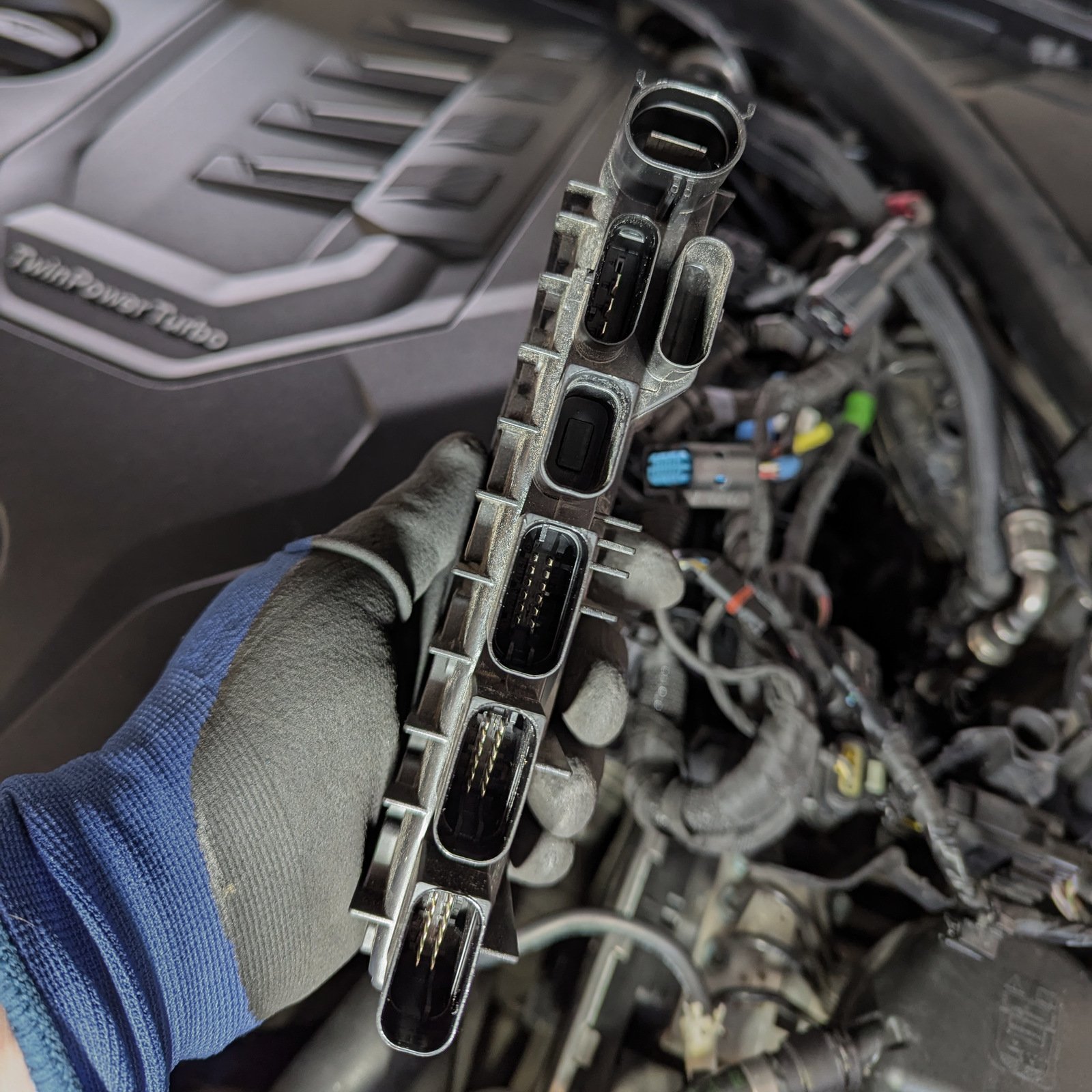 Electronic control module held in a gloved hand beside a BMW engine bay.